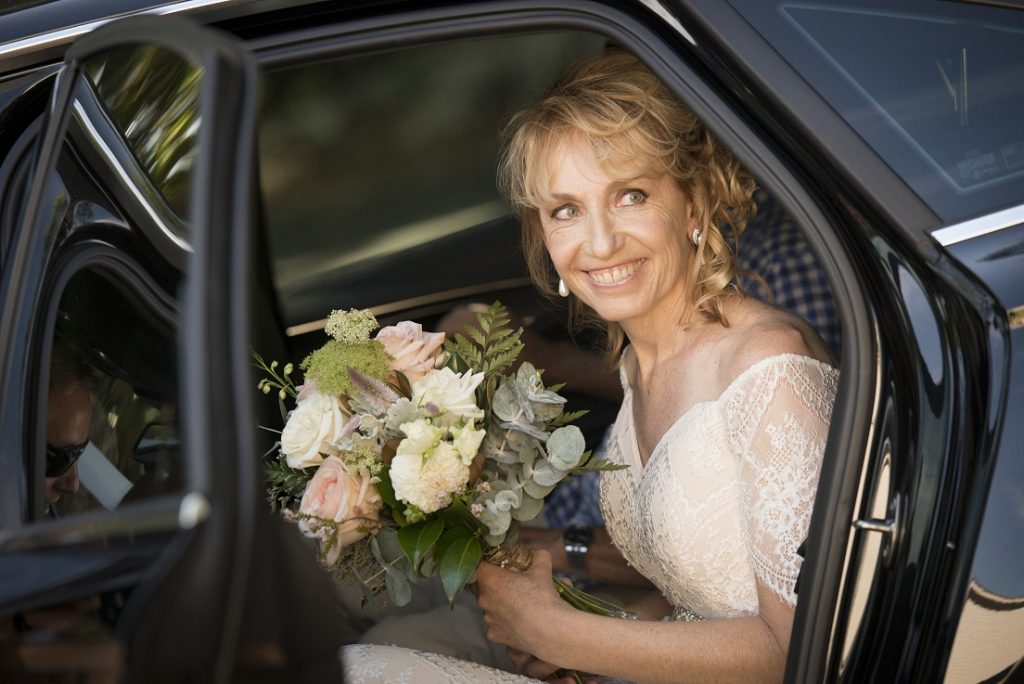Woman wearing a wedding dress & smiling while seated in a bridal car holding a bouquet of flowers