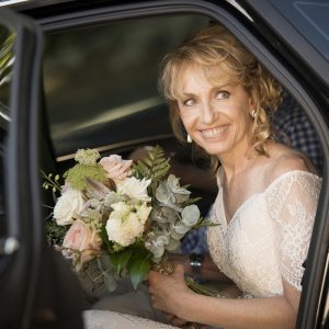 Woman wearing a wedding dress & smiling while seated in a bridal car holding a bouquet of flowers